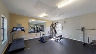 This fitness center is a dedicated, single-floor exercise facility designed to support a well-rounded workout routine. The space is finished with durable dark rubber flooring throughout, providing a safe and functional surface ideal for a range of physical activities. The room is bathed in natural light from multiple windows, including a set of elegant French doors that open to an outdoor area, creating an airy and motivating atmosphere. Overhead fluorescent lighting ensures the space remains well-lit at all times.

The facility is outfitted with a variety of equipment to accommodate diverse fitness needs. Cardiovascular options include a treadmill and a stationary bike, while strength training is supported by a multi-function cable machine and a barbell rack with a weight bench for free weight exercises. A large wall-mounted mirror spans a significant portion of one wall, enhancing the sense of space and allowing users to monitor their form during workouts. A wall-mounted fan provides additional ventilation and comfort. Safety is addressed with a clearly visible fire extinguisher on the wall. The neutral-toned walls and clean, uncluttered layout contribute to a focused and functional environment suited for daily fitness use.