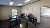 This single-floor community amenity space is thoughtfully designed to serve as both a business center and a casual gathering area. The room features a dedicated workstation area along one wall, where a granite-topped desk accommodates multiple computer terminals with ergonomic seating — ideal for residents needing a productive workspace. Decorative circular metal wall art adds a contemporary touch to the neutral beige walls, creating a welcoming atmosphere.

The space is divided into distinct functional zones, with multiple round bistro-style tables and black high-back chairs arranged throughout, providing comfortable spots for informal meetings or social interaction. A popcorn machine and a media console with a printer add practical and recreational amenities to the room.

Patterned carpeting in dark tones runs throughout, complementing the dark furnishings and crown molding that frames the room's perimeter. Flush-mounted fluorescent ceiling fixtures provide consistent, bright overhead lighting. Multiple windows fitted with white horizontal blinds allow natural light to filter in while offering views of the surrounding outdoor greenery, contributing to an open and airy feel. The overall ambiance is polished yet approachable, blending functionality with comfort.
