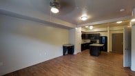 This single-floor space offers an open-concept design that seamlessly connects the living area and kitchen, creating a cohesive and functional layout. Warm hardwood-style flooring flows throughout, adding a sense of continuity and comfort to the space. The living area is illuminated by a ceiling fan with integrated lighting, providing both airflow and ambient light.

The kitchen is a standout feature, showcasing rich dark cabinetry that contrasts elegantly with light granite-style countertops. A central island provides additional prep space and serves as a natural gathering point. Black appliances, including a refrigerator, microwave, and range, complement the cabinetry for a cohesive, modern aesthetic. A bar-style counter with additional dark cabinetry bridges the kitchen and living areas, offering extra storage and serving functionality.

The textured ceiling adds subtle visual interest overhead, while flush-mount lighting fixtures ensure the kitchen remains bright and well-lit. Neutral wall tones throughout keep the space feeling open and versatile, ready to accommodate a variety of interior styles. Overall, the space combines practical design with modern finishes to deliver a well-appointed, inviting environment.