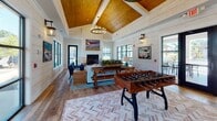 This single-story space showcases a thoughtfully designed interior that seamlessly combines rustic farmhouse character with modern finishes. Throughout the open layout, white-painted shiplap walls create a clean, airy backdrop, while warm wood-paneled vaulted ceilings with exposed beams add architectural depth and a sense of grandeur. The flooring transitions between wide-plank reclaimed hardwood and a distinctive herringbone-pattern brick tile, grounding the space with rich texture and visual interest.

A central lounge area anchors the space, highlighted by a whitewashed brick fireplace with a rustic wood mantle and wall-mounted television above. Comfortable seating arranged around a wood coffee table invites relaxation, while black-framed picture windows flood the area with natural light and provide views to an outdoor patio. A foosball table nearby adds a recreational element, making the space equally suited for leisure and entertainment.

The wet bar area features deep green cabinetry with brass hardware, a light quartz countertop, open floating wood shelves, and stainless steel appliances. A dining area with sturdy wood tables and woven rattan chairs provides ample seating, completing this well-rounded, inviting gathering space.