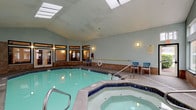 This single-level indoor aquatic facility is designed with both functionality and comfort in mind. The centerpiece is a generously sized swimming pool with clear, aqua-tinted water, bordered by cobalt blue tile accents and equipped with stainless steel entry rails for easy access. Adjacent to the pool sits a circular hot tub, similarly finished with blue tile detailing, providing a dedicated relaxation area.

The vaulted ceiling features multiple skylights that draw in abundant natural light, complementing the soft sage green walls and warm brown stone tile wainscoting that lines the perimeter. Large, multi-pane windows and glass-paneled doors line one side of the facility, offering views of the surrounding greenery and allowing additional daylight to filter through.

The pool deck is finished in a textured aggregate surface, providing a slip-resistant walkway around the water. Lightweight seating chairs in blue and white are arranged throughout the space, offering convenient poolside rest areas. Wall-mounted sconce lighting adds warmth during low-light hours. Safety signage, a life ring, and a handrail along the pool edge reflect the facility's attention to safety standards throughout.