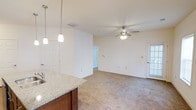 This single-floor space showcases a seamless open-concept layout connecting the kitchen and living area. The kitchen features a granite countertop peninsula with rich dark wood cabinetry and stylish pendant lighting that hangs overhead, providing both functional task lighting and a decorative focal point. The peninsula serves as a natural divider between the kitchen and the living area, creating a sense of separation while maintaining an open, airy feel.

The living area is finished with plush, neutral-toned carpeting that extends throughout, adding warmth and comfort underfoot. A ceiling fan with integrated lighting is centrally positioned, offering both air circulation and ambient illumination. The walls are finished in a clean, neutral tone, complementing the overall brightness of the space.

Natural light enters through a windowed door on the far wall, enhancing the openness of the room. A doorway provides access to additional areas of the home. The combination of practical finishes, thoughtful lighting, and an open floor plan creates a functional and welcoming everyday living environment.