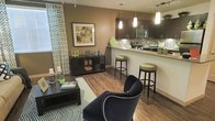 This single-floor space showcases a thoughtfully designed open-concept layout that effortlessly connects the living area and kitchen into one cohesive, functional environment. The living area features rich hardwood-style flooring, a geometric-patterned area rug, and warm taupe walls that create an inviting atmosphere. A bold navy accent chair with nailhead trim serves as a striking focal point, complemented by a dark wood media console and coordinating coffee table. Bold patterned curtains frame the window, adding a lively visual contrast to the neutral walls. The kitchen is defined by dark espresso cabinetry, a mosaic tile backsplash, and sleek stainless steel appliances. A peninsula with light granite countertops bridges the two spaces, offering bar-height seating with green upholstered stools that echo the space's accent color palette. Pendant lighting above the peninsula and track lighting overhead provide layered illumination throughout. The open flow between the kitchen and living area makes the space feel expansive and well-connected, ideal for both everyday living and entertaining. The cohesive color scheme of warm browns, deep navy, and fresh green accents gives the space a polished, modern character.