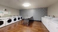 Located at 1400 W Third St in Chico, California, this shared laundry facility is a well-appointed, single-floor space designed to serve the practical needs of its users efficiently and comfortably. The room is generously sized, offering ample open floor space that allows for easy movement throughout.

The most visually striking element is the bold black-and-white geometric tile wall, featuring a repeating pinwheel pattern that spans a full accent wall and wraps into an adjacent corner, giving the space a modern, design-forward character. This statement wall is complemented by warm wood-look flooring that runs throughout, creating a clean and inviting contrast.

Multiple front-loading washing machines and top-loading dryers line the walls, providing substantial shared laundry capacity. A stainless steel utility sink with a prep table sits near the entry, offering a convenient handwashing and pre-treatment station. A central stainless steel folding table on casters provides a practical surface for sorting and folding laundry.

Overhead, a flush-mount ceiling light fixture provides bright, even illumination across the room. A ventilation soffit above the sink area supports proper airflow. The space is accessed through a glass-paneled door and also includes a wall-mounted fire extinguisher, underscoring attention to safety. Overall, the facility is clean, functional, and thoughtfully finished.