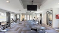 Located at 3251 Overton Rd in Birmingham, Alabama, this well-appointed fitness center offers a comprehensive workout environment spread across a single floor. The space is designed with a clean, modern aesthetic, featuring crisp white walls, recessed lighting, and a distinctive herringbone-patterned carpet in neutral tones of gray and tan that runs throughout the entire facility.

The fitness center is generously proportioned, with an elongated open floor plan that allows for a diverse array of exercise equipment. Cardio machines, including treadmills by Star Trac Pro and various elliptical trainers, are strategically positioned to take advantage of the large windows that line the walls, offering views of the surrounding greenery. A graceful arched window anchors one end of the room, flooding the treadmill area with natural light.

Strength training equipment, including a multi-station cable machine and adjustable weight benches, occupies one section of the space. Large floor-to-ceiling mirrors are mounted along the walls to enhance the sense of openness and assist with proper form. Ceiling fans and a ceiling-mounted flat-screen television add comfort and entertainment during workouts.

French doors with glass panels open to the exterior, further connecting the interior to the outdoors. An adjacent area visible through a half-wall opening suggests access to a lounge or common area, rounding out the amenities of this thoughtfully designed fitness facility.