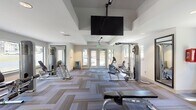 Located at 3251 Overton Rd in Birmingham, Alabama, this well-appointed fitness center offers a comprehensive workout environment spread across a single floor. The space is designed with a clean, modern aesthetic, featuring crisp white walls, recessed lighting, and a distinctive herringbone-patterned carpet in neutral tones of gray and tan that runs throughout the entire facility.

The fitness center is generously proportioned, with an elongated open floor plan that allows for a diverse array of exercise equipment. Cardio machines, including treadmills by Star Trac Pro and various elliptical trainers, are strategically positioned to take advantage of the large windows that line the walls, offering views of the surrounding greenery. A graceful arched window anchors one end of the room, flooding the treadmill area with natural light.

Strength training equipment, including a multi-station cable machine and adjustable weight benches, occupies one section of the space. Large floor-to-ceiling mirrors are mounted along the walls to enhance the sense of openness and assist with proper form. Ceiling fans and a ceiling-mounted flat-screen television add comfort and entertainment during workouts.

French doors with glass panels open to the exterior, further connecting the interior to the outdoors. An adjacent area visible through a half-wall opening suggests access to a lounge or common area, rounding out the amenities of this thoughtfully designed fitness facility.