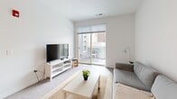 Located at 101 Main St in Lafayette, Indiana, this well-designed one-bedroom, one-bathroom unit offers a thoughtful layout across a single floor. The open-concept kitchen and living area serves as the heart of the space, where a large peninsula with a white quartz countertop and built-in sink doubles as a breakfast bar with modern bar stools. Pendant lighting with glass cylinder shades adds a contemporary touch overhead, while dark espresso cabinetry provides a rich contrast. Stainless steel appliances, including a refrigerator, range, and microwave, complete the fully equipped kitchen. A stacked washer and dryer unit is conveniently integrated nearby. The living area flows naturally toward a sliding glass door that opens onto a private balcony, allowing natural light to fill the room. The bedroom offers a calm, neutral atmosphere with carpeted flooring, soft wall tones, and a window fitted with blinds for light control. The bathroom features a dark wood vanity with ample drawer storage, a white countertop, an undermount sink, and well-placed wall sconces flanking a large mirror. A dedicated closet provides additional storage, rounding out this efficiently designed and modern urban residence.