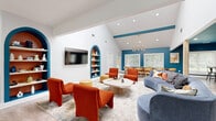 Located at 7150 W Peachtree Dunwoody Rd NE in Atlanta, GA, this single-floor community space delivers a well-appointed, open-concept layout designed for both social gatherings and everyday use. Light oak-toned hardwood flooring runs continuously throughout, grounding the space with warmth and cohesion.

The living and lounge area is the centerpiece, featuring soaring vaulted ceilings accented by a bold teal-painted beam and an eye-catching sputnik chandelier. Vibrant orange lounge chairs, a curved slate-blue sofa, and a round coffee table are arranged around an area rug, creating an inviting conversational setting. Flanking built-in arched shelving units, framed in teal with terracotta-painted interiors, add a striking architectural detail and display decorative accents.

A large conference-style dining table with leather chairs occupies an adjacent zone, ideal for meetings or communal dining. Natural light pours through multiple large windows offering views of surrounding greenery. A vertical slatted wood partition subtly delineates functional zones without closing off the open feel.

The kitchen is clean and functional, outfitted with white shaker cabinetry, subway tile backsplash, dual stainless steel French door refrigerators, and built-in microwaves — well-equipped to support group events. The entry welcomes visitors through full-height glass doors, reinforcing the space's bright and accessible character.