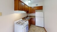 This single-floor unit is thoughtfully laid out to maximize functionality across its key living areas. The main living space features warm hardwood-style flooring that extends throughout, complemented by white walls and wood-trimmed doorways that lend a consistent, cohesive aesthetic. Natural light filters in through multiple windows fitted with horizontal blinds, brightening the interior throughout the day. A louvered closet door adds a classic detail to the living area, which also provides direct access to an exterior door.

The kitchen is efficiently designed with warm wood cabinetry wrapping around two walls, offering ample upper and lower storage. Dark granite-style countertops provide a practical work surface, while white appliances — including a gas range and a full-size refrigerator — complete the space. Tile flooring runs underfoot, and a ventilation hood is positioned above the range.

The bedroom is carpeted in a neutral tone and includes a ceiling fan with an integrated light fixture, ensuring year-round comfort. A window allows natural light to enter the room.

The bathroom is accessed through a wood-paneled door and features tile flooring, a combined tub-shower with a curtain rod, and a window above the tub for added ventilation and light. The overall unit presents a functional and practical living environment.