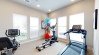 This dedicated fitness space is thoughtfully designed to support a comprehensive workout experience within a single, open room. The interior features neutral beige walls and light carpet flooring, creating a clean and inviting environment conducive to exercise. Recessed lighting is complemented by a ceiling fan, ensuring the space remains well-lit and comfortable during use.

Natural light is a defining characteristic of this room, with multiple large windows fitted with white horizontal blinds lining the walls and providing views of the surrounding exterior. A large wall-mounted mirror on one side amplifies the sense of space and allows for form-checking during strength training.

The cardio zone is well-appointed, housing a treadmill, elliptical trainer, rowing machine, and a recumbent stationary bike—all Spirit-branded equipment. A multi-functional cable machine with an attached weight stack and adjustable bench occupies the strength training area, alongside a dumbbell rack stocked with a range of free weights. Exercise balls and framed athletic photography add functional and aesthetic touches. A wall-mounted flat-screen TV provides entertainment during workouts, rounding out this fully functional fitness space.