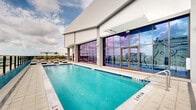 Located at 307 SW 5th St in Fort Lauderdale, this rooftop amenity level delivers a resort-caliber outdoor experience high above the city skyline. The centerpiece is a generously sized swimming pool with vivid turquoise water, framed by clean-lined tile coping and floor-to-ceiling glass walls that connect the pool terrace to an adjacent indoor space. A separate spa offers an intimate relaxation zone, accented by a bold teal accent wall that adds a vibrant pop of color to the modern aesthetic. The expansive sun deck is outfitted with an array of striped black-and-white chaise lounges and shaded cabana areas, providing ample space for sunbathing and leisure. Glass railing systems along the perimeter ensure unobstructed, panoramic views of the Fort Lauderdale skyline, stretching across the surrounding urban landscape. Tropical landscaping, including palm trees and lush greenery in raised planters, introduces a natural, Florida-inspired ambiance to the space. An interior lounge area features large glass sliding doors that open onto the deck, showcasing vibrant tropical mural artwork and creating a seamless indoor-outdoor connection. The overall design combines contemporary architecture with a lively, South Florida aesthetic throughout.