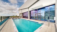 Located at 307 SW 5th St in Fort Lauderdale, this rooftop amenity level delivers a resort-caliber outdoor experience high above the city skyline. The centerpiece is a generously sized swimming pool with vivid turquoise water, framed by clean-lined tile coping and floor-to-ceiling glass walls that connect the pool terrace to an adjacent indoor space. A separate spa offers an intimate relaxation zone, accented by a bold teal accent wall that adds a vibrant pop of color to the modern aesthetic. The expansive sun deck is outfitted with an array of striped black-and-white chaise lounges and shaded cabana areas, providing ample space for sunbathing and leisure. Glass railing systems along the perimeter ensure unobstructed, panoramic views of the Fort Lauderdale skyline, stretching across the surrounding urban landscape. Tropical landscaping, including palm trees and lush greenery in raised planters, introduces a natural, Florida-inspired ambiance to the space. An interior lounge area features large glass sliding doors that open onto the deck, showcasing vibrant tropical mural artwork and creating a seamless indoor-outdoor connection. The overall design combines contemporary architecture with a lively, South Florida aesthetic throughout.