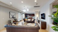 This single-floor space is designed as an open, multifunctional lounge environment that seamlessly integrates several distinct functional zones within one continuous layout. Light hardwood flooring runs throughout, lending warmth and visual cohesion to the entire space.

The main seating area is anchored by tufted leather Chesterfield sofas arranged around a pair of glass-and-metal coffee tables, creating an inviting and stylish focal point. Globe pendant lights hang overhead, complementing recessed ceiling lighting to provide a well-lit atmosphere. A large wall-mounted flat-screen television is positioned above a modern media console with frosted glass cabinet doors, offering an entertainment-focused area alongside the lounge seating.

Natural light filters through multiple windows fitted with horizontal blinds along one wall, enhancing the bright, airy feel of the space. A dedicated workstation area with bar-height seating and a desk setup is positioned near the windows, making it a functional zone for focused work.

The kitchenette is fully equipped with rich dark wood cabinetry, granite countertops, a mosaic tile backsplash, stainless steel appliances including a refrigerator and microwave, and a single-basin sink. A standalone beverage station adds further convenience. Artwork and greenery are thoughtfully placed throughout, adding personality and life to the overall environment.