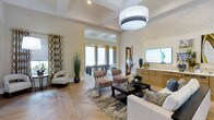 This single-story residence showcases an open-concept layout that seamlessly connects a refined main living area to an adjoining game room, creating a generous and versatile entertaining space. The living area is anchored by a striking textured stone accent wall, which frames a large flat-screen television mounted above a warm natural wood media console. A substantial drum pendant light hangs overhead, complementing the coffered ceiling detail that adds architectural depth to the room.

The flooring throughout is a standout feature — light-toned hardwood laid in a classic herringbone pattern adds visual interest and elegance underfoot. Seating is arranged around a dark-stained coffee table atop a patterned area rug, creating an inviting conversation zone. A pair of upholstered accent chairs and stools provide additional flexible seating.

The adjoining game room, visible through a wide open archway, benefits from an abundance of natural light streaming through large windows, illuminating a billiards table and additional seating. Patterned drapery panels frame the windows, adding warmth and texture. Together, the rooms offer a cohesive, well-appointed space ideal for both relaxed living and entertaining.