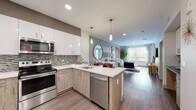This contemporary single-floor residence in Walnut Creek, California offers a thoughtfully designed open-concept layout with modern finishes throughout. The kitchen is a standout feature, equipped with stainless steel appliances including a range, microwave, dishwasher, and refrigerator. Two-tone cabinetry pairs white upper cabinets with wood-grain lower cabinets, complemented by a mosaic tile backsplash and crisp white quartz countertops. A peninsula with seating provides additional prep space and connects seamlessly to the dining area, where a dedicated bar-height table with pendant lighting creates a stylish entertaining zone. The open living area is bathed in natural light from large windows that frame outdoor greenery, creating a bright and welcoming ambiance. Wood-style flooring runs throughout the main living spaces, adding warmth and continuity. The bedroom is carpeted and features a bold accent wall, large windows with outdoor views, and generous proportions. The bathroom offers a wood-grain vanity with white countertop and a large mirror, and connects directly to the bedroom. A hallway utility closet houses a full-size stacked washer and dryer for added convenience. A second bathroom with a bold red accent wall adds a vibrant design touch. Recessed lighting throughout enhances the modern aesthetic of this well-appointed home.