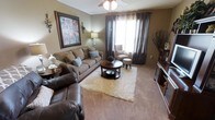 This single-floor residence offers a welcoming and cohesive interior designed with comfort and style in mind. The living room serves as the heart of the home, showcasing a neutral color palette of warm taupes and earthy browns that create a relaxed yet polished ambiance. Plush wall-to-wall carpeting adds softness underfoot, complemented by a layered area rug that anchors the seating arrangement. The room is furnished with a generously sized upholstered sofa accented with decorative pillows, a leather recliner, and a cushioned accent chair paired with an ottoman, creating ample seating for relaxing or entertaining. A round wooden coffee table serves as a central focal point, while a matching side table adds functional charm. Natural light filters in through large windows dressed with elegant floor-length drape curtains, brightening the space. A ceiling fan with integrated lighting provides both comfort and illumination. Decorative wall sconces and a framed piece of wall art add character and a touch of sophistication. A wooden shelving unit along one wall offers additional display and storage. The overall atmosphere is warm, lived-in, and thoughtfully curated, making for a comfortable and inviting single-level home.