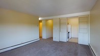 This single-floor space offers an open, functional layout that integrates the living area and kitchen within one cohesive environment. The living area is finished with wall-to-wall carpet in a neutral gray tone, providing a soft, understated foundation. Warm yellow-toned walls contrast with crisp white trim and structural columns, lending the space a clean yet inviting character. A baseboard heating unit runs along the exterior wall, ensuring practical climate control. Built-in open shelving is integrated into one of the white dividing columns, offering convenient storage and display space. The kitchen area, partially visible beyond the dividing columns, features tile flooring, a white refrigerator, and a range, providing essential appliances in a compact, efficient footprint. A hallway leading to additional areas of the space is accessible from the main living zone, suggesting further utility beyond what is immediately visible. The ceiling is smooth and white, contributing to a bright, airy feel. Overall, the space presents a straightforward, practical design with a neutral palette that provides a versatile and move-in ready environment.