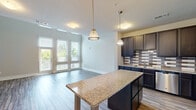 This two-story space is designed with a modern aesthetic and thoughtful layout spread across two levels. The ground floor hosts an open-plan kitchen and living area, where wood-look flooring flows seamlessly throughout. The kitchen is well-appointed with dark espresso cabinetry, a geometric tile backsplash, stainless steel appliances, and a granite-topped center island illuminated by pendant lighting. The living area benefits from floor-to-ceiling windows and a glass-paneled door that floods the space with natural light and outdoor views. A striking architectural feature is the double-height ceiling in the living area, adding a dramatic sense of volume and openness.

A modern staircase with a sleek white railing connects to the upper level, which offers a carpeted bedroom area with built-in shelving, a walk-in closet, and an in-unit washer. Both bathrooms are finished with dark vanity cabinetry, white countertops, framed mirrors, and tile flooring, maintaining a consistent design language throughout. The overall ambiance is contemporary and bright, with a neutral color palette enhancing the sense of spaciousness across both floors.