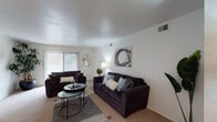 This single-story residence offers a thoughtfully designed layout that balances comfort and functionality across its key living spaces. The living room serves as the central gathering area, finished with neutral-toned carpet and a textured ceiling. A full-width sliding glass door fitted with vertical blinds fills the room with natural light and provides direct access to an outdoor patio, offering pleasant exterior views. The space accommodates two dark upholstered sofas arranged around a tiered glass coffee table, creating a comfortable and cohesive seating arrangement.

Adjacent to the living room, the dining area features carpet flooring and a ceiling fan with integrated lighting. An open passage connects this space directly to the kitchen, which is equipped with rich dark wood cabinetry, dark granite-style countertops, a double basin sink, and black appliances including a refrigerator and dishwasher. Wood-look flooring adds warmth to the kitchen.

The bathroom is finished with matching dark wood cabinetry, a single vanity with a dark countertop, a large mirror, and a tub-shower combination. Two bedrooms, each carpeted and simply appointed, offer quiet, private retreats with ample wall space and soft, neutral color palettes throughout.