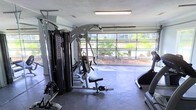 This single-floor fitness space is purpose-built for serious training, offering a well-equipped environment for a comprehensive workout routine. The centerpiece of the room is a robust multi-station cable and weight machine, providing a wide range of strength-training exercises in one compact unit. A cardio machine — visible along the far wall — complements the strength equipment, making this a well-rounded training facility.

The flooring is fitted with durable rubber gym mats, providing cushioning and traction suited for intense exercise. Overhead fluorescent lighting ensures the space is well-lit at all hours, while the room's most striking architectural feature — a full-width glass-panel garage door — invites an abundance of natural light and offers a pleasant view of the surrounding outdoor landscape, including palm trees and neighboring greenery. This feature also allows for excellent ventilation when opened, creating a refreshing, open-air workout experience.

The walls provide ample vertical storage space, with fitness accessories and attachments organized along one side. The overall ambiance is functional and focused, with a clean layout that maximizes usable floor space while maintaining an uncluttered environment ideal for an effective and motivating fitness routine.