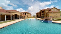 This residential community clubhouse showcases a beautifully designed outdoor amenity space centered around a large, resort-style swimming pool with crystal-clear blue water. The pool area is a standout feature, complete with a decorative water fountain wall finished in mosaic tile, providing an elegant focal point. Surrounding the pool is a generous concrete pool deck with lounge chairs, all enclosed by wrought iron fencing that adds both security and a refined aesthetic.

The covered patio adjacent to the pool area is supported by classic white columns with arched detailing, creating a shaded retreat with comfortable seating. Large gridded windows and glass doors connect the interior clubhouse to the outdoor space, offering unobstructed pool views from within. The exterior is finished in a warm stucco with tile roofing, consistent with the Mediterranean-inspired architectural style seen throughout the surrounding multi-story residential buildings.

Manicured lawns, concrete walkways, and mature palm trees complete the landscaping, contributing to an open and inviting atmosphere. The community's design emphasizes indoor-outdoor connectivity, with the clubhouse serving as a central hub overlooking the pool and green spaces. The overall ambiance is bright, airy, and well-suited for communal living and leisure activities.