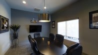 This single-floor professional meeting space is thoughtfully designed to facilitate productive collaboration in a refined, intimate setting. The centerpiece of the room is a large, round dark conference table surrounded by comfortable black leather executive chairs, comfortably seating a small group for focused discussions or presentations. A bold drum-style metallic pendant light hangs overhead, casting warm, direct illumination over the table and adding a touch of contemporary elegance to the space. The walls are finished in a sophisticated muted gray tone, complementing the dark furnishings and creating a calm, professional atmosphere. A wall-mounted flat-screen display provides a convenient visual aid for presentations and meetings, positioned prominently for easy viewing from all seats. Framed artwork and branded décor add personality and a sense of organizational identity to the room. A large window fitted with a roller shade allows natural light to filter into the space while maintaining privacy, offering a bright and airy feel. The carpeted flooring adds acoustic comfort, making the environment well-suited for conversation. Overall, this conference room strikes a balance between functionality and style, making it an ideal venue for business meetings, strategic planning sessions, or team gatherings.