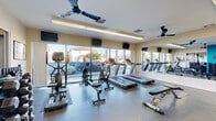 This fitness center in National City, California, is a thoughtfully designed, single-floor facility offering a comprehensive range of exercise equipment to support both cardio and strength training. The space is divided into distinct functional zones, creating an organized and efficient workout environment.

The cardio area is anchored by a bank of floor-to-ceiling windows that flood the room with natural light and provide open views of the surrounding neighborhood. Treadmills, cycling bikes, and elliptical-style machines are arranged along this windowed wall, each positioned in front of wall-mounted televisions for entertainment during workouts. Ceiling fans circulate air throughout the space, contributing to a comfortable exercise environment.

A large wall mirror spans one side of the room, visually expanding the space and allowing users to monitor their form. The strength training section features a multi-function cable machine, a dumbbell rack with a full range of free weights, kettlebells, and an adjustable workout bench. A dedicated area with a teal accent wall adds a vibrant visual element and separates workout zones.

The flooring throughout consists of durable, textured rubber tiles suited for high-traffic fitness use. The overall ambiance is clean, bright, and functional, making this facility a well-rounded option for residents seeking convenient access to fitness amenities.