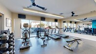 This fitness center in National City, California, is a thoughtfully designed, single-floor facility offering a comprehensive range of exercise equipment to support both cardio and strength training. The space is divided into distinct functional zones, creating an organized and efficient workout environment.

The cardio area is anchored by a bank of floor-to-ceiling windows that flood the room with natural light and provide open views of the surrounding neighborhood. Treadmills, cycling bikes, and elliptical-style machines are arranged along this windowed wall, each positioned in front of wall-mounted televisions for entertainment during workouts. Ceiling fans circulate air throughout the space, contributing to a comfortable exercise environment.

A large wall mirror spans one side of the room, visually expanding the space and allowing users to monitor their form. The strength training section features a multi-function cable machine, a dumbbell rack with a full range of free weights, kettlebells, and an adjustable workout bench. A dedicated area with a teal accent wall adds a vibrant visual element and separates workout zones.

The flooring throughout consists of durable, textured rubber tiles suited for high-traffic fitness use. The overall ambiance is clean, bright, and functional, making this facility a well-rounded option for residents seeking convenient access to fitness amenities.