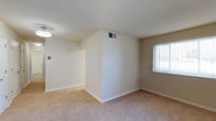 This single-floor space presents a clean, functional interior characterized by neutral beige walls and wall-to-wall carpeting in a warm, light tone. The open layout allows for a seamless visual connection between the main room and an adjacent area, creating a sense of continuity throughout the space. A central dividing wall adds architectural definition while maintaining an open feel. Natural light enters through a vertical blind-covered window, brightening the interior and complementing the light color palette. HVAC vents are positioned in the ceiling and on the wall, indicating a central climate control system. White baseboards line the perimeter, providing a clean contrast against the beige walls and carpet. Electrical outlets are conveniently placed throughout, offering practical utility. The overall ambiance is simple and understated, with a move-in ready condition that provides a neutral canvas for personalization. The space benefits from a straightforward, efficient layout suited for everyday living.