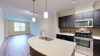 Located at 12190 Waveland St in Fairfax, Virginia, this thoughtfully designed one-bedroom, one-bathroom residence offers a comfortable and modern living experience. The open-concept kitchen and living area serves as the heart of the home, anchored by a curved peninsula island with a stainless steel sink and quartz-style countertops. Dark espresso cabinetry with sleek hardware lines the kitchen walls, complemented by a tile backsplash, stainless steel appliances including a refrigerator, range, and microwave, and elegant crown molding overhead. Pendant lighting and a track light fixture illuminate the space, while wood-look flooring flows throughout the main living area, adding warmth and continuity. The living room benefits from natural light through a large window. The bedroom is a generously sized retreat finished with plush carpet and a window overlooking the surrounding neighborhood. An expansive walk-in closet fitted with multi-tier wire shelving provides excellent storage. The bathroom features a soaking tub with a tiled surround and neutral finishes. Crown molding throughout the main living areas adds a refined architectural touch, tying the entire space together with a polished, cohesive aesthetic.