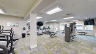 Located in Hillsboro, Oregon, this fitness center offers a spacious, single-floor exercise room thoughtfully designed to accommodate a full range of workout activities. The room is generously proportioned, providing ample space for both cardio and strength training without feeling cramped.

The flooring features a distinctive modular carpet tile pattern in shades of green, gray, and yellow, lending a modern, energetic aesthetic to the space. Recessed skylight-style flush ceiling panels provide bright, even illumination throughout, complemented by natural light streaming in through multiple windows that offer views of the surrounding greenery.

The cardio section is equipped with a row of TRUE brand treadmills and multiple elliptical trainers, each featuring integrated consoles. Wall-mounted flat-screen televisions are positioned throughout the room for entertainment during workouts. The strength training area includes a multi-function cable machine, a leg press machine, an adjustable weight bench, and additional resistance equipment, offering users a comprehensive range of training options.

Full-length mirrors line one wall, visually expanding the space and allowing users to monitor their form. A water station is conveniently accessible within the room. The overall design is clean and contemporary, with light green and white walls, white structural columns, and a well-organized layout that ensures smooth circulation throughout the fitness center.