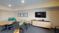 Located in Ramsey, Minnesota, this single-floor community space is designed to accommodate a variety of social and recreational activities in a welcoming, open environment. The layout is generously proportioned, with distinct zones that flow seamlessly into one another across carpeted and hard-surface flooring.

The fully equipped kitchen serves as a central hub, featuring dark espresso cabinetry, dark countertops, a black refrigerator, range, dishwasher, and microwave. A built-in island with bar-height seating provides additional prep and gathering space, while industrial-style pendant lights suspended from a track system add purposeful task lighting and visual character. Warm yellow-toned walls carry throughout the space, contributing to an inviting atmosphere.

Adjacent to the kitchen, multiple dining tables with plaid-upholstered and mixed seating chairs accommodate groups of varying sizes. A dedicated lounge area is furnished with green accent chairs and features a large wall-mounted flat-screen television flanked by framed artwork, creating a comfortable media and relaxation zone.

A bookshelf stocked with games and reading materials occupies one corner near a secondary seating cluster, offering quiet leisure options. Natural light filters in through a window along one wall, and a secure exterior entry door provides convenient direct access to the space.