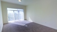 This single-floor space offers a clean and functional layout with a light, airy atmosphere. The main living area features wall-to-wall dark carpeting that adds warmth underfoot, complemented by smooth, neutral-toned walls that create a calm and versatile backdrop. A large sliding glass door serves as a prominent architectural feature, framed by vertical blinds that allow for easy light control. When open, the blinds permit generous streams of natural light to flood the interior, casting bright patterns across the floor and enhancing the sense of openness. The flush-mount ceiling light provides additional illumination, ensuring the space remains well-lit at any time of day. The room's simple, uncluttered design makes it highly adaptable for a variety of uses and furniture arrangements. The sliding glass door hints at an outdoor area beyond, offering the possibility of a patio, balcony, or exterior access. Overall, the space presents a straightforward, well-maintained environment with practical finishes and a welcoming natural brightness that defines its overall character.