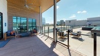 Located in Fort Worth, Texas, this contemporary rooftop terrace offers a sophisticated outdoor retreat designed for both relaxation and social gatherings. The space seamlessly blends covered and open-air sections, connected by large floor-to-ceiling glass walls and sliding doors that flood the interior with natural light while maintaining a visual connection to the surrounding cityscape.

The covered lounge area is anchored by a striking wood-paneled accent wall, upon which a wall-mounted flat-screen television is centered for outdoor entertainment. A woven rattan pendant light hangs above, adding a warm, organic touch to the modern design. Comfortable navy blue seating with wood frames and burnt orange accent pillows is arranged around a low-profile coffee table, creating an inviting social setting. The ceiling overhead is finished in warm wood tones, complementing the textured wall cladding.

The open-air portion of the terrace features wide-plank composite decking and is outfitted with additional lounge seating, a ceiling fan for warm Texas days, and glass panel railings that preserve unobstructed panoramic views of the Fort Worth skyline. The overall design balances clean, modern architecture with natural materials, resulting in an elevated outdoor space that is both functional and visually compelling.