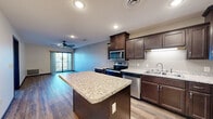 Located at 212 Locust St in Monticello, MN, this three-bedroom, one-bathroom unit offers a well-designed single-floor layout that maximizes functionality across its compact footprint. The open-concept kitchen and living area serves as the heart of the home, showcasing rich dark-walnut cabinetry, granite-look countertops, and a full suite of stainless steel appliances including a range, microwave, dishwasher, and refrigerator. A central kitchen island provides additional prep and dining space. Wood-look plank flooring runs throughout the main living areas, adding warmth and visual continuity. The living room opens directly to a private balcony through a sliding glass door, bringing in natural light and outdoor views of the surrounding neighborhood. The three bedrooms are carpeted for comfort, with the spacious primary bedroom featuring a walk-in closet area adjacent to the full bathroom. The bathroom is finished with dark wood cabinetry, a vanity, and wood-look plank flooring consistent with the rest of the unit. Secondary bedrooms are well-proportioned with neutral walls and adequate natural light. The overall aesthetic is clean and modern, with neutral tones and consistent finishes throughout, creating a cohesive and move-in-ready living environment.