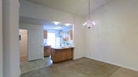 This single-floor space features an open-concept design that seamlessly connects the living and dining areas to the kitchen. The dining area is anchored by a delicate candelabra-style chandelier, adding a touch of classic elegance to the otherwise neutral, understated space. Neutral-toned carpet covers the living and dining areas, contributing to a warm and cohesive feel throughout.

The kitchen is accessible through an open passageway and centers around a functional peninsula with a light-colored countertop, offering additional workspace and a casual seating or serving area. Warm wood-toned cabinetry lines both the upper and lower sections of the kitchen, providing ample storage. White appliances, including a refrigerator and range, complement the cabinetry. A window above the sink allows natural light to brighten the kitchen, while a fluorescent overhead fixture provides additional illumination. A laundry area is also visible, adding convenience to the layout. The overall ambiance is simple, functional, and move-in ready.