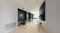 This contemporary single-floor space is thoughtfully designed to serve as a professional business center or coworking lounge. The interior is defined by a crisp white palette with polished tile flooring that reflects natural light throughout, creating a bright and open atmosphere.

A standout feature is the row of semi-private work alcoves lining one side of the space. Each alcove is framed by deep, dark-toned cabinetry and rich patterned fabric panels, with individual pendant lighting overhead and a dedicated work surface — offering focused, booth-like workspaces within the open floor plan. Along the opposite wall, a wall-mounted desk surface with monitor mounts provides additional workstation options.

A dedicated print and copy station is recessed into a vibrant teal mosaic tile niche, paired with a built-in white counter and lower cabinetry, adding a bold accent to the otherwise neutral space. Sheer curtains at a nearby window allow diffused natural light to filter in softly.

The lounge area features a generous navy sectional sofa accented with teal throw pillows, complemented by contemporary dining-style seating and dark wood tables. Colorful geometric artwork adorns the walls, adding energy to the space. Floor-to-ceiling glass windows and a glass door on one end open to views of a landscaped outdoor area and surrounding streetscape, seamlessly connecting the interior to the outside environment.
