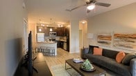 This single-floor residence showcases a thoughtfully designed open-concept layout that integrates the kitchen and living area into one cohesive, functional space. The kitchen is a standout feature, outfitted with rich dark wood cabinetry, stainless steel appliances including a refrigerator, microwave, and range, as well as a built-in sink. A curved breakfast bar with barstool seating provides a casual dining option and serves as a natural dividing point between the kitchen and the rest of the living space. Pendant lighting and a track lighting system overhead illuminate the kitchen with warm, layered light, enhancing the ambiance of the dark cabinetry and light stone countertops. The flooring throughout is light-toned hardwood, lending a clean and airy feel to the space. The living area is comfortably appointed with a contemporary sofa and a low-profile coffee table, anchored by a soft area rug. A ceiling fan centered above provides both comfort and an additional source of ambient lighting. Neutral wall tones complement the warm wood and modern furnishings, creating an inviting atmosphere. A hallway visible from the main space suggests additional rooms beyond, contributing to the overall sense of a well-organized and efficiently planned floor plan.