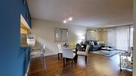 This single-floor space showcases an open-concept design that integrates the dining and living areas into one cohesive, flowing layout. Rich, warm-toned hardwood flooring extends throughout, anchoring the space and adding a sense of continuity. The neutral beige walls provide a versatile backdrop that complements the contemporary furnishings and accent colors of yellow and blue visible throughout.

The dining area features a round glass-top table surrounded by upholstered white chairs with nailhead trim detailing, positioned beneath track lighting mounted on the ceiling. A pair of chrome-framed mirrors adorn the wall, adding depth and a modern decorative touch. Adjacent to the dining area, a breakfast bar with white bar stools offers additional seating.

The living area is anchored by a plush gray sectional sofa accented with patterned throw pillows. Patterned drapery frames the background, softening the space and adding visual interest. A textured area rug defines the seating zone. The textured ceiling adds subtle dimension overhead, while the overall ambiance is warm, well-lit, and inviting throughout.