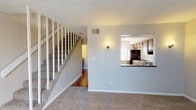 Located at 218 Olympia Dr in Homewood, Alabama, this two-story residence offers a practical and comfortable layout spread across two well-connected levels. The main living area is generously sized, finished with neutral-toned carpet and bathed in natural light through a large sliding glass door that opens to a private balcony. Vertical blinds provide shade and privacy while maintaining the bright, open feel of the space. A white-railed staircase connects the two floors, serving as a clean architectural feature within the living area. The kitchen is positioned adjacent to the living room and features a convenient pass-through window, warm wood cabinetry, dark laminate countertops, and black appliances including a range, dishwasher, refrigerator, and built-in microwave. Hardwood-style flooring runs through the kitchen and bathroom areas, providing a durable contrast to the plush carpet found in the living and bedroom spaces. The bathroom includes a full tub-shower combination and connects directly to an adjacent bedroom, which benefits from its own window allowing natural light to filter in. Throughout the home, neutral gray walls, textured ceilings, and consistent finishes create a cohesive and understated aesthetic suited for a variety of living arrangements.