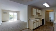 This single-floor space offers a practical and comfortable layout with an open-concept design that connects the kitchen and living area seamlessly. The kitchen is equipped with light wood cabinetry providing ample storage, a dual-basin sink, and tile flooring that transitions into the carpeted living space. The cabinetry extends to the upper walls, maximizing vertical storage capacity. A built-in dishwasher and extended countertop workspace add to the kitchen's functionality. The living area features wall-to-wall carpeting, lending a warm and inviting atmosphere to the space. A textured ceiling adds subtle character throughout. The standout feature of the living area is the full-width sliding glass door that opens to a private balcony, flooding the interior with natural light and offering pleasant views of the surrounding landscape. The overall layout is efficient and well-organized, with a clear visual connection between the kitchen and living zones, making the space feel open and airy. Neutral tones throughout the unit provide a clean, versatile backdrop suitable for a variety of interior styles.