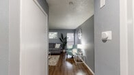 This single-story space is designed with a consistent aesthetic, featuring cool gray walls, wood-look flooring, and textured ceilings throughout. A central hallway connects the main living areas, offering an immediate view into the living room upon entry.

The living room serves as the heart of the space, accommodating a sofa, accent chair, and media console. Two windows allow natural light to filter in, while a wall-mounted mirror adds depth to the room. A decorative wall clock and tall potted plant contribute to the overall character of the space.

The kitchen features dark marbled countertops that contrast with white cabinetry equipped with brushed nickel hardware. Upper cabinets provide storage space, and under-cabinet lighting illuminates the work surface. White appliances, including a refrigerator and dishwasher, complete the kitchen.

The bedroom is a calm retreat with gray walls, textured ceiling, and wood-look flooring. A large sliding glass door with vertical blinds allows natural light and outdoor views. The room includes closet access.

The bathroom mirrors the kitchen's aesthetic with a matching dark marbled countertop, an undermount sink, and a large wall mirror. Built-in side cabinetry provides additional storage in this well-appointed space.