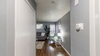 This single-story space is designed with a consistent aesthetic, featuring cool gray walls, wood-look flooring, and textured ceilings throughout. A central hallway connects the main living areas, offering an immediate view into the living room upon entry.

The living room serves as the heart of the space, accommodating a sofa, accent chair, and media console. Two windows allow natural light to filter in, while a wall-mounted mirror adds depth to the room. A decorative wall clock and tall potted plant contribute to the overall character of the space.

The kitchen features dark marbled countertops that contrast with white cabinetry equipped with brushed nickel hardware. Upper cabinets provide storage space, and under-cabinet lighting illuminates the work surface. White appliances, including a refrigerator and dishwasher, complete the kitchen.

The bedroom is a calm retreat with gray walls, textured ceiling, and wood-look flooring. A large sliding glass door with vertical blinds allows natural light and outdoor views. The room includes closet access.

The bathroom mirrors the kitchen's aesthetic with a matching dark marbled countertop, an undermount sink, and a large wall mirror. Built-in side cabinetry provides additional storage in this well-appointed space.