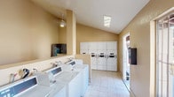 Located at 633 Ascot in Vista, this community laundry facility is a practical, well-organized space designed to accommodate the needs of its residents. The single-floor layout features warm beige walls and light tile flooring throughout, creating a clean and functional environment. Vaulted ceilings add a sense of openness to the room, and the space is illuminated by a combination of recessed skylight-style flush fixtures and pendant lighting.

The facility is equipped with a row of commercial-grade top-loading washing machines, each featuring digital controls for ease of use. Opposite the washers, a bank of stacked commercial dryers lines the wall, numbered for easy identification and offering ample drying capacity. A granite-style folding counter runs along one wall, providing generous surface space for sorting and folding laundry. An additional wall-mounted shelf extends the available workspace in a corner section of the room.

Natural light filters in through a barred window, offering a view to the exterior. A vending machine is conveniently located near the entrance, providing easy access to laundry supplies. The facility also features an exterior-facing door, allowing for straightforward access from the surrounding community. Overall, this laundry facility is thoughtfully equipped to serve its residents efficiently and comfortably.