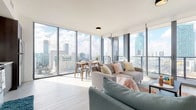 Unit 1908 is a thoughtfully designed high-rise residence that combines contemporary architecture with stunning panoramic city views. The open-concept living and dining area is bathed in natural light, thanks to expansive floor-to-ceiling windows that wrap around the corner of the unit, offering sweeping skyline vistas from every angle. Light-toned hardwood-style flooring runs throughout, enhancing the bright, airy atmosphere of the space.

The kitchen is outfitted with sleek, dark wood cabinetry, white countertops, and a full suite of stainless steel appliances, including a range, microwave, and refrigerator — providing both style and full functionality.

The unit features two well-appointed bedrooms. The primary bedroom enjoys large floor-to-ceiling windows framing impressive city views, while the second bedroom offers a quieter, more intimate setting with access to a closet. Both rooms feature the same light flooring that carries through the rest of the residence.

Two bathrooms serve the unit. The primary bathroom includes a glass-enclosed walk-in shower with large-format tile work and modern fixtures. The second bathroom features a vanity with ample storage and clean, contemporary finishes.

Overall, Unit 1908 delivers a refined urban living experience, defined by its luminous interiors, high-end finishes, and unobstructed city skyline views.