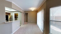 This single-floor residence offers a clean, functional layout designed for comfortable everyday living. The main living area is finished with soft, wall-to-wall carpeting in a neutral tone, creating a warm and inviting atmosphere throughout. Crown molding lines the ceiling, adding a refined architectural detail that elevates the overall aesthetic of the space.

A standout feature of the living area is the expansive floor-to-ceiling vertical blind-covered sliding glass doors, which allow an abundance of natural light to pour in while offering direct access to the outdoors. The neutral taupe wall color complements the light-toned carpet and white trim, creating a cohesive and understated color palette.

Adjacent to the living area, a peninsula counter with a granite surface defines the boundary of the kitchen, which is visible through an open pass-through. The kitchen includes built-in shelving and cabinetry, offering practical storage solutions. A hallway leading from the living space provides access to additional rooms, with a utility or storage closet visible along the corridor. Flush-mount ceiling lighting provides consistent illumination throughout, and ample electrical outlets are positioned conveniently along the baseboards. Overall, the space presents a tidy, move-in-ready environment with a balanced blend of practicality and classic design elements.