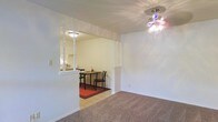 This single-floor residence offers a thoughtfully arranged layout that balances openness with defined functional areas. The living area features wall-to-wall carpeting that adds warmth underfoot, while a decorative chandelier with colorful blue and clear glass accents serves as a striking focal point overhead, casting a distinctive light pattern across the white ceiling.

A structural beam with decorative columns creates a natural transition between the living area and the dining space, providing architectural definition without fully enclosing the rooms. The dining area is finished with light tile flooring, contrasting with the carpeted living space and grounded by a rich red area rug beneath a simple dining table and chairs set.

A pass-through opening in the wall connects the dining area to the kitchen, enhancing the sense of flow and openness throughout the home. The kitchen is visible through this opening, hinting at its functional layout. Throughout the space, clean white walls provide a neutral, bright backdrop that enhances the sense of openness. The overall ambiance is cozy yet practical, with design elements that create clear distinctions between living, dining, and kitchen zones while maintaining a cohesive, connected feel across the single floor.