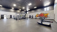 The Elliott Premier Apartment Homes Fitness Center in Kannapolis, NC, is a generously sized, single-floor facility designed to accommodate a wide range of fitness activities. The space is organized into distinct zones, each with its own character and purpose.

The main workout hall features high ceilings, exposed concrete block walls, and durable rubber flooring throughout. This industrial-style area is energized by bold, large-scale graffiti murals — including a striking Hulk mural and vibrant typographic art — creating a motivating, urban atmosphere. The space is equipped with a multi-functional power rack, a full dumbbell rack, cable machines, battle ropes, sleds, and various strength training stations, providing comprehensive options for serious training.

A secondary, elegantly appointed studio offers a stark contrast in aesthetic. Featuring soaring coffered ceilings, oversized arched windows that flood the room with natural light, and views of the surrounding landscape, this bright and airy space houses spin bikes, a rowing machine, resistance equipment, stability balls, and a ballet barre with mirrored wall — ideal for cardio and group fitness activities.

The entrance foyer is welcoming, with polished concrete floors, arched architectural detailing, bold double glass doors, and prominent Elliott branding. The facility seamlessly blends industrial energy with refined architectural elements to serve a diverse range of fitness needs.
