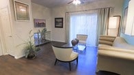 This single-floor residence offers a thoughtfully designed living space that blends elegance with comfort. The living room serves as the centerpiece of the home, showcasing rich dark hardwood flooring that contrasts beautifully with the light-toned walls adorned with classic crown molding. A ceiling fan with a decorative light fixture adds both style and practicality to the space. Large sliding glass doors, framed by floor-to-ceiling vertical blinds and elegant striped drapes, flood the room with natural light and provide a seamless connection to the outdoors. The living area is furnished with a curated mix of contemporary and classic pieces, including plush neutral-toned seating, a sleek glass coffee table, and an accent console table with decorative vases. Framed artwork on the walls adds a personal touch and enhances the refined ambiance. A lush indoor plant introduces a refreshing natural element to the interior. The overall atmosphere is bright, airy, and well-appointed, creating an inviting environment that is both functional and aesthetically pleasing. The cohesive design palette of warm neutrals and dark wood tones lends the space a sophisticated yet welcoming character throughout.