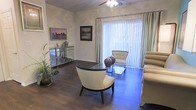 This single-floor residence offers a thoughtfully designed living space that blends elegance with comfort. The living room serves as the centerpiece of the home, showcasing rich dark hardwood flooring that contrasts beautifully with the light-toned walls adorned with classic crown molding. A ceiling fan with a decorative light fixture adds both style and practicality to the space. Large sliding glass doors, framed by floor-to-ceiling vertical blinds and elegant striped drapes, flood the room with natural light and provide a seamless connection to the outdoors. The living area is furnished with a curated mix of contemporary and classic pieces, including plush neutral-toned seating, a sleek glass coffee table, and an accent console table with decorative vases. Framed artwork on the walls adds a personal touch and enhances the refined ambiance. A lush indoor plant introduces a refreshing natural element to the interior. The overall atmosphere is bright, airy, and well-appointed, creating an inviting environment that is both functional and aesthetically pleasing. The cohesive design palette of warm neutrals and dark wood tones lends the space a sophisticated yet welcoming character throughout.